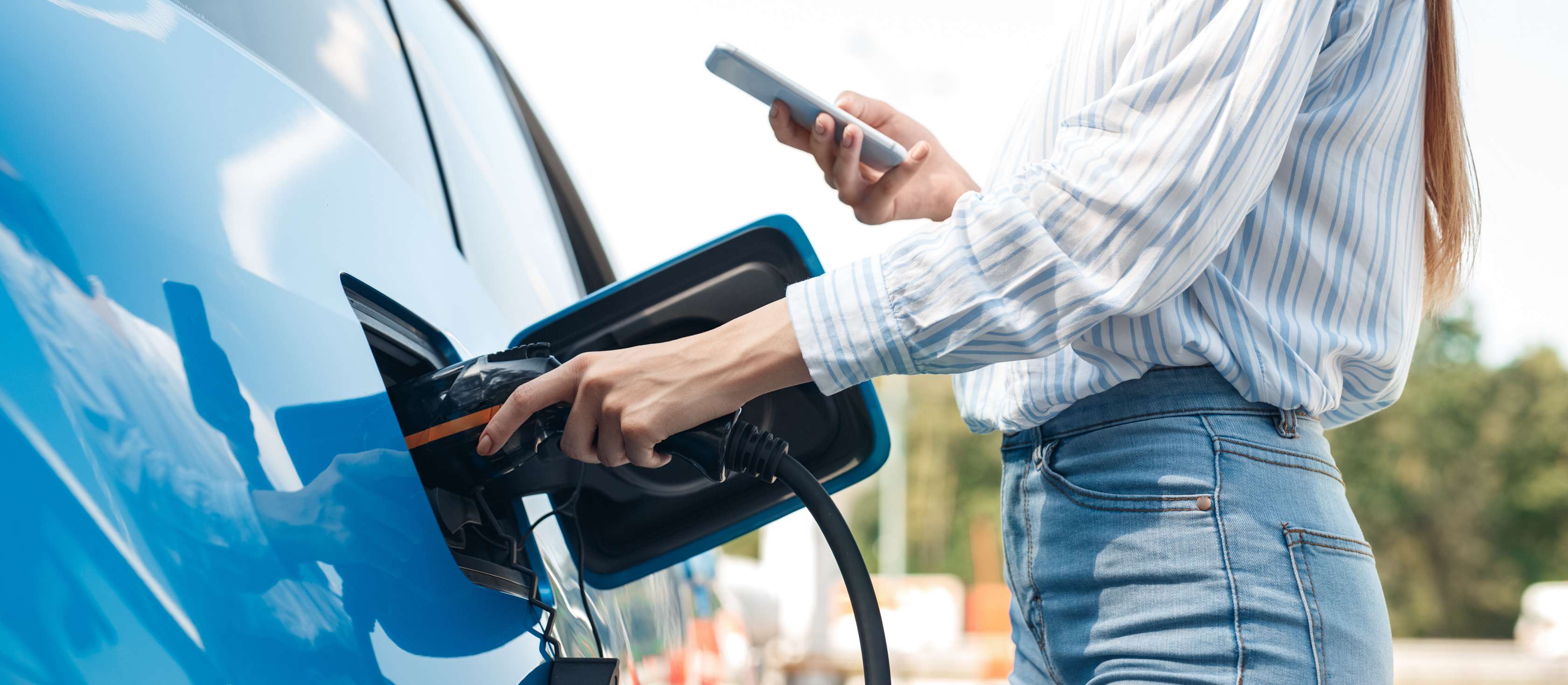 E-Auto laden Versicherung: Frau mit Smartphone in der Hand befestigt Ladekabel an Ladebuchse eines blauen E-Autos