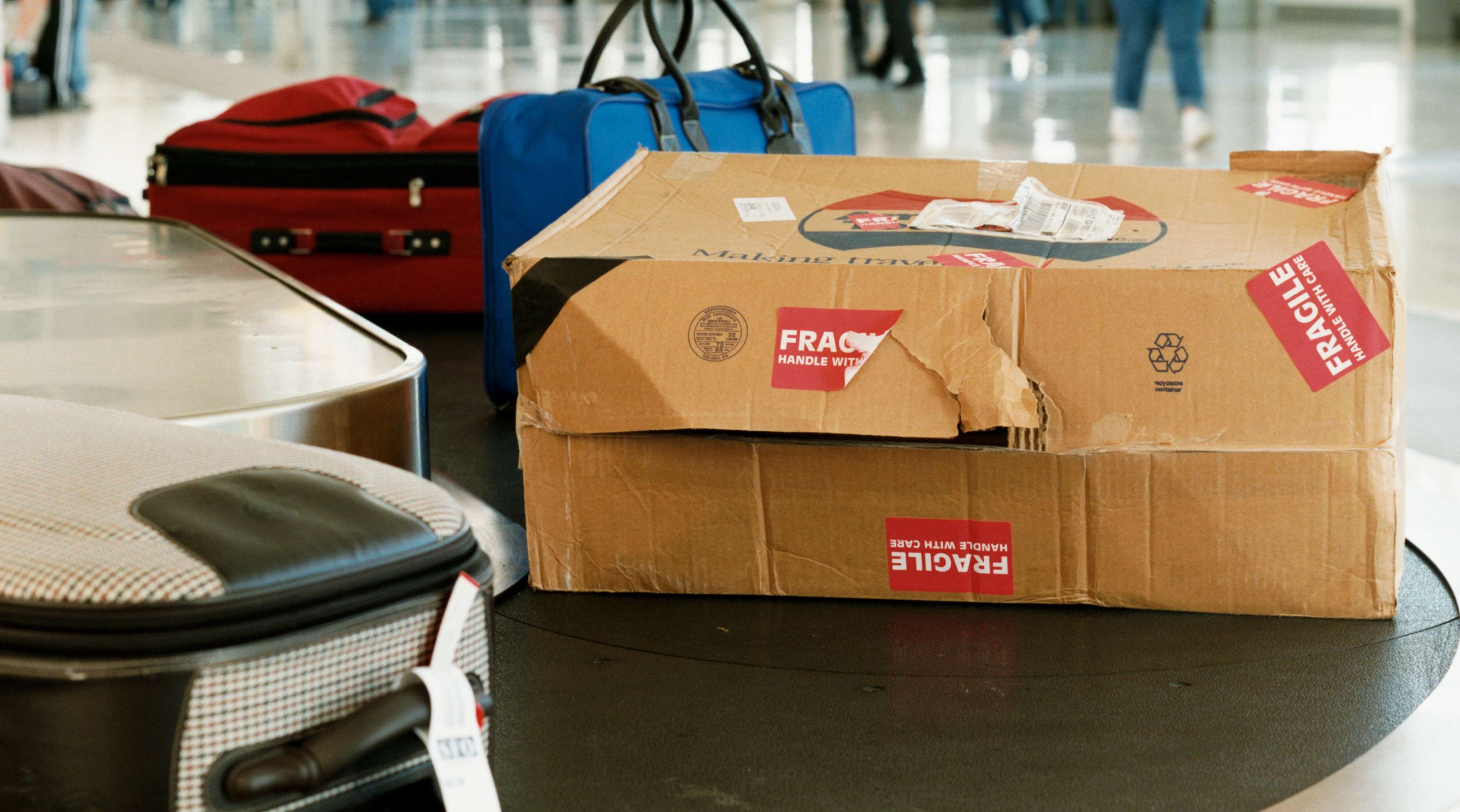 Allianz - Beschädigtes Reisegepäck: Koffer und Pappkarton auf Gepäckförderband