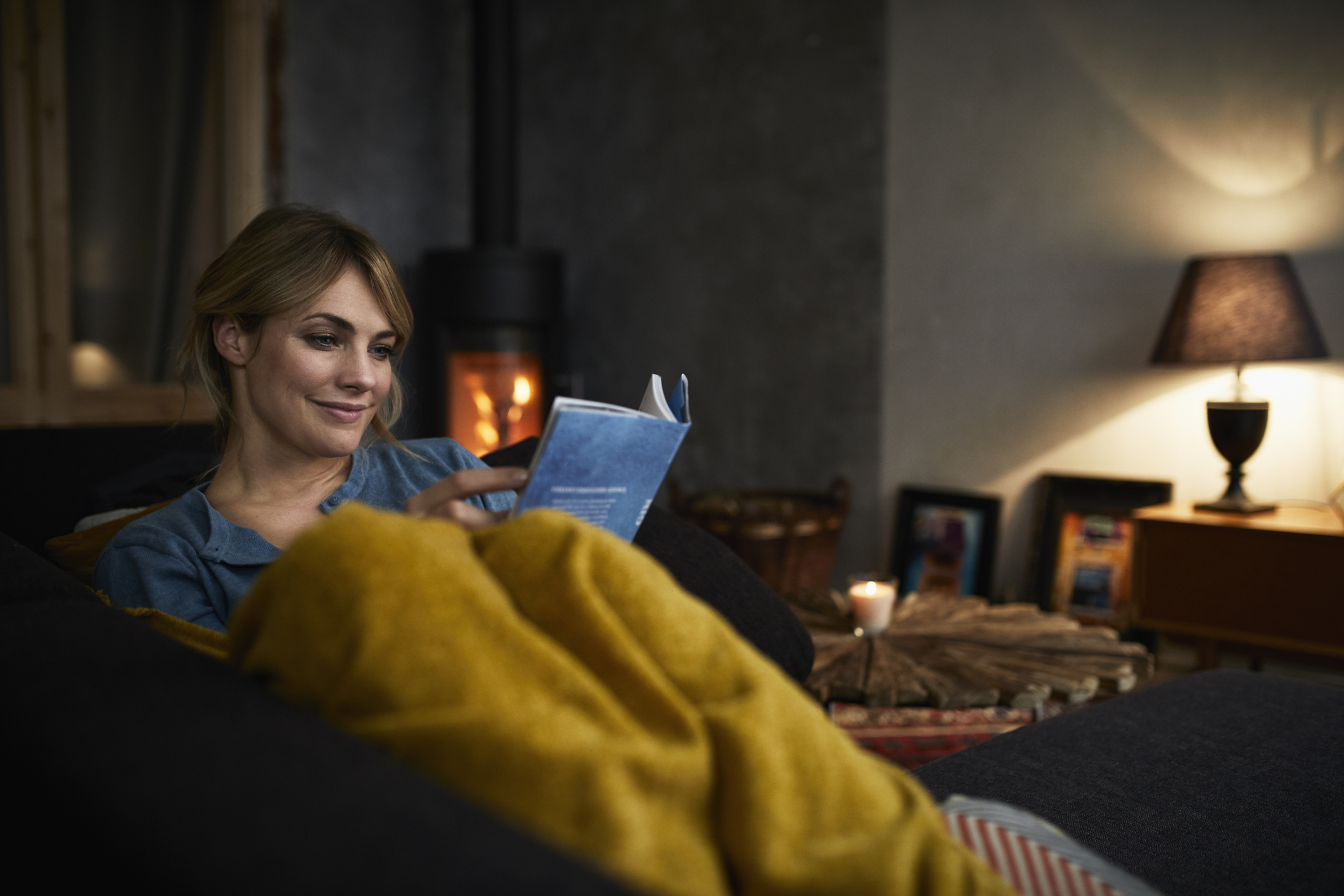 Frau sitzt in eine Decke gehüllt in Wohnzimmer und liest ein Buch
