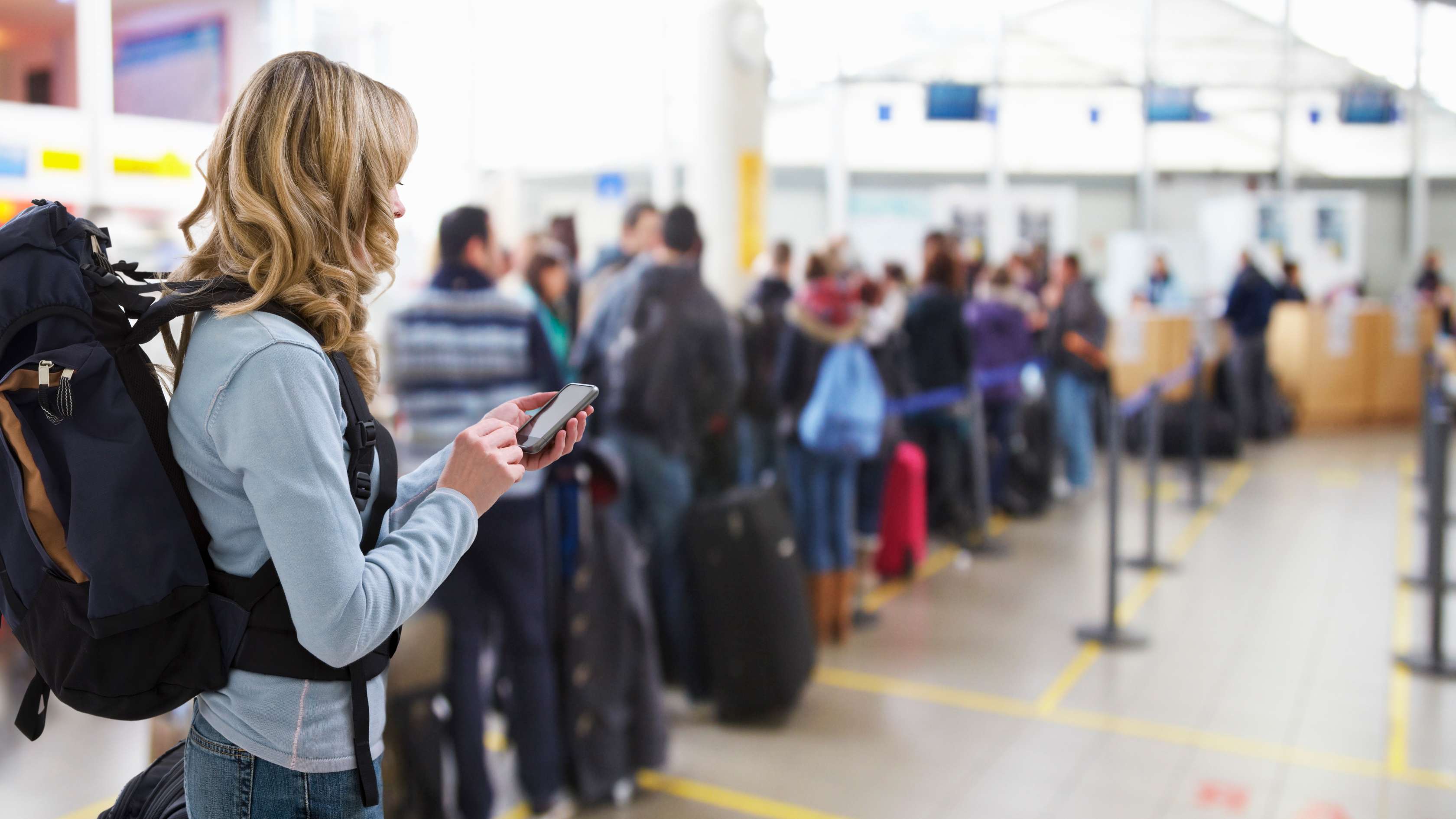 Frau mit Rucksack auf dem Rücken steht in einer langen Schlange am Flughafenschalter und sieht auf ihr Handy.