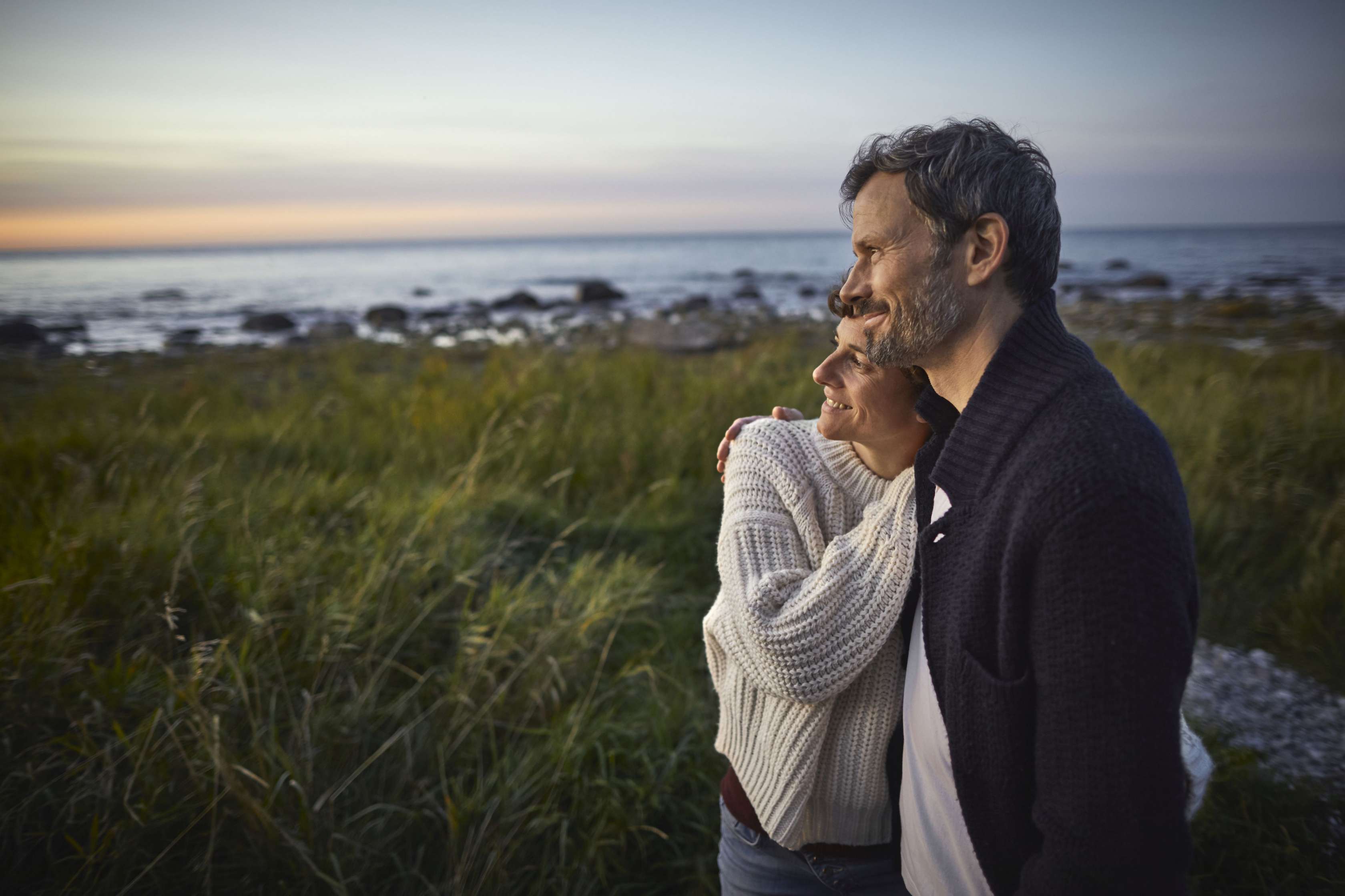 Allianz - Allianz Reiserücktrittsversicherung: Lächelndes Paar blickt Arm in Arm aufs Meer hinaus